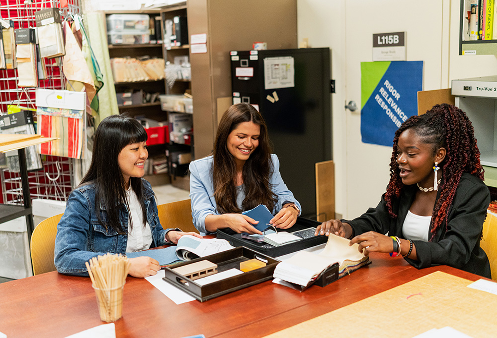 Three students sit at a table smiling. Various material samples and design tools sit on the table.